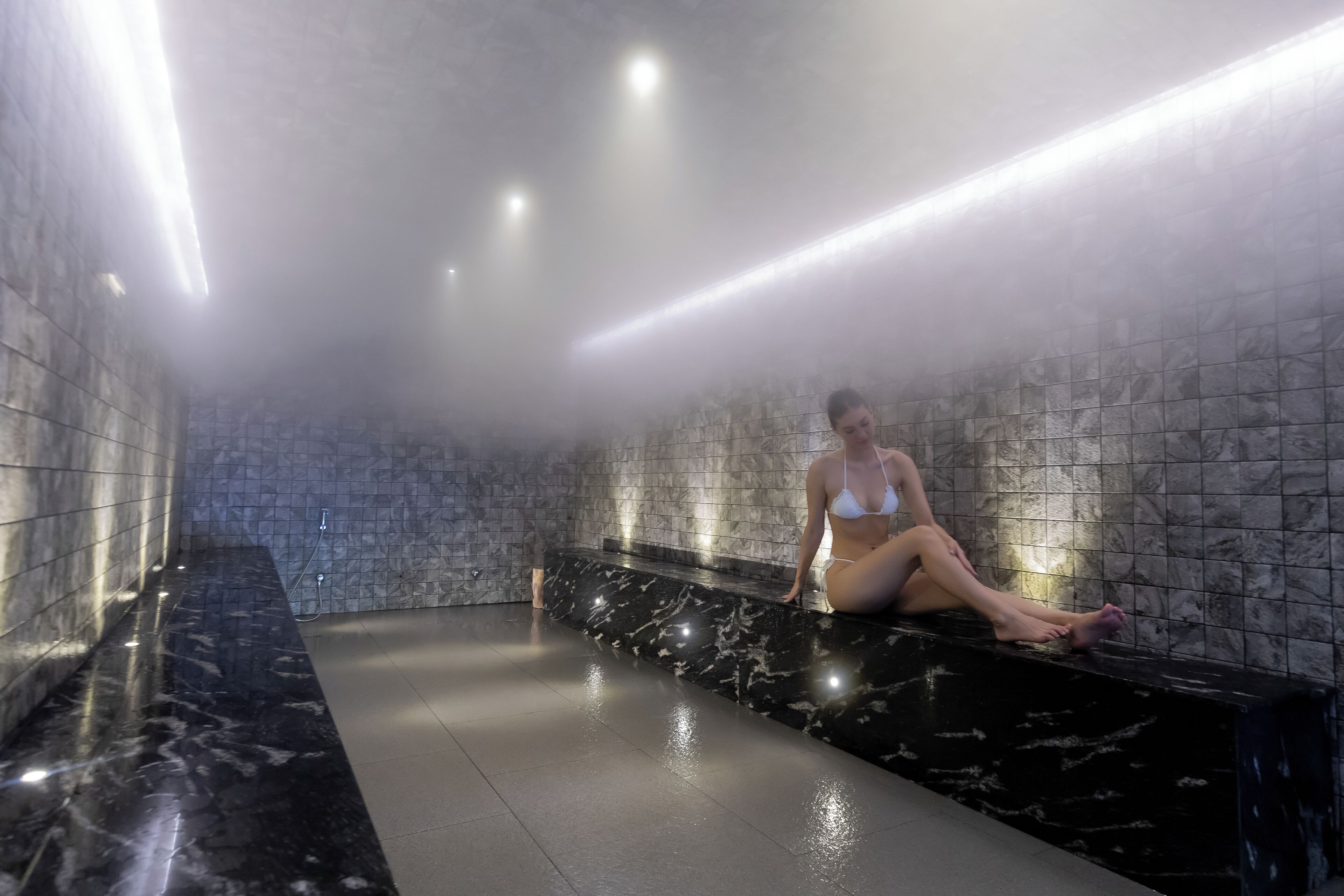 A person sitting on a black marble bench in a steam room with tiled walls and a tiled floor.
