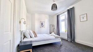 A bedroom with a bed, bedside table, wall-mounted lamp, and a window with curtains. - Moorland House by Switchback Stays (Cardiff)