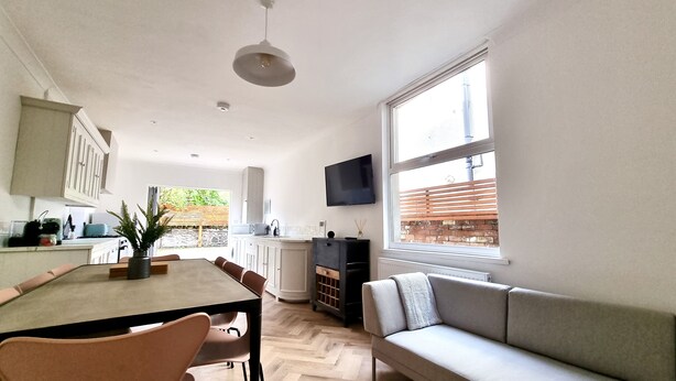 A modern kitchen with white cabinets, a dining table with chairs, a built-in oven, and a large window with a view of greenery. - Moorland House by Switchback Stays (Cardiff)