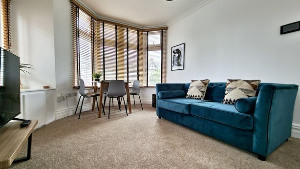 Una sala de estar con un sofá azul, dos sillones, una mesa de centro y un televisor. Hay ventanas grandes con persianas, un cuadro en la pared y una planta en el alféizar.