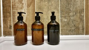 Tres botellas de champú, acondicionador y gel de baño sobre una superficie blanca con fondo de madera.