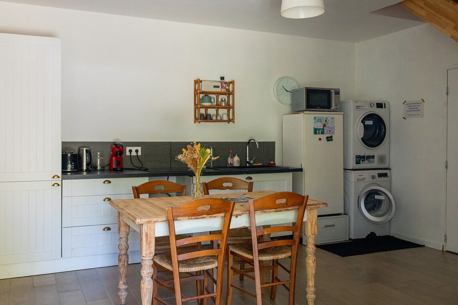 A compact kitchen with a dining table, chairs, a microwave, a washing machine, and a clock on the wall.