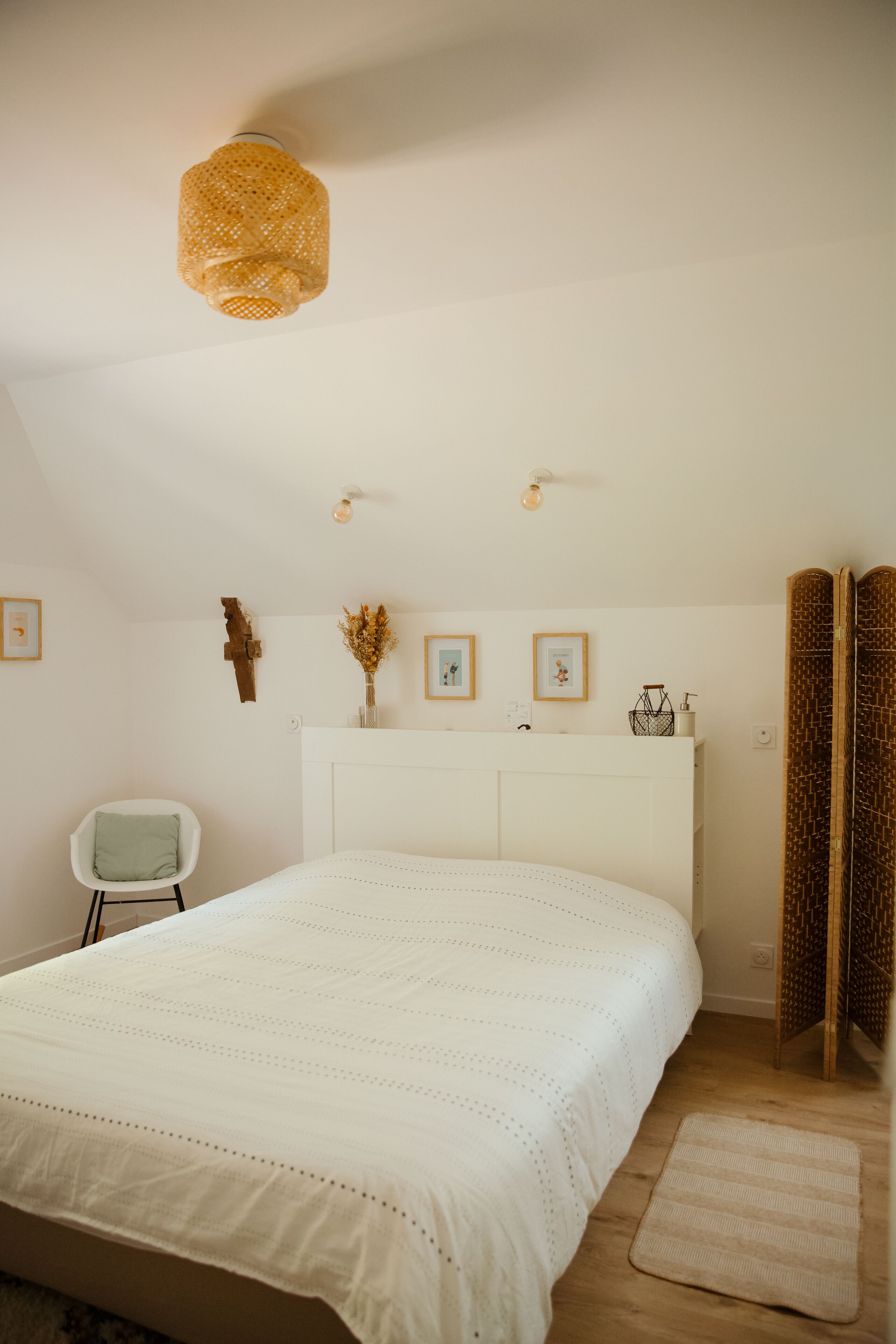 Une chambre à coucher avec un grand lit, une chaise blanche, une table de chevet avec un vase et un luminaire fixé au mur.