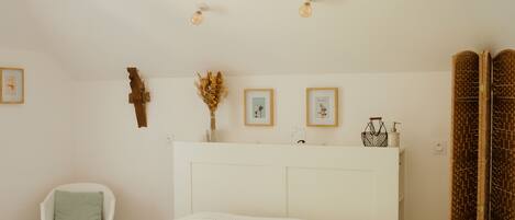 Une chambre à coucher avec un grand lit, une chaise blanche, une table de chevet avec un vase et un luminaire fixé au mur.