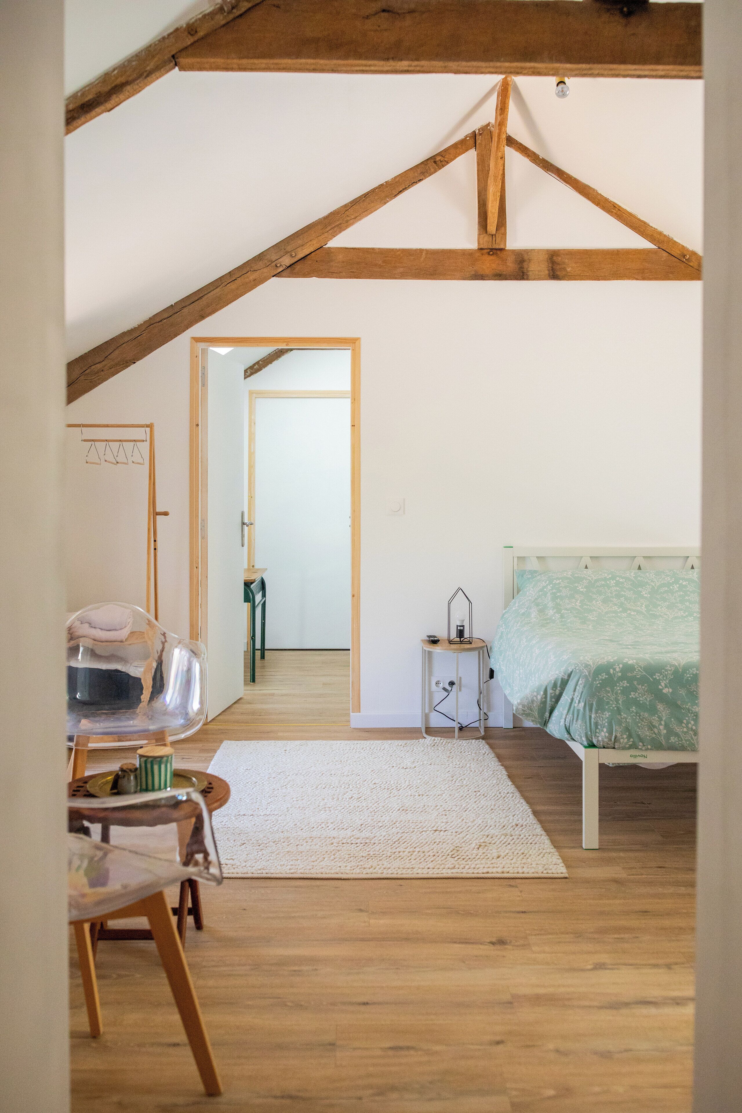 A bedroom with a wooden ceiling, a bed with a green floral bedspread, a bedside table, and a chair with a clear plastic cover.