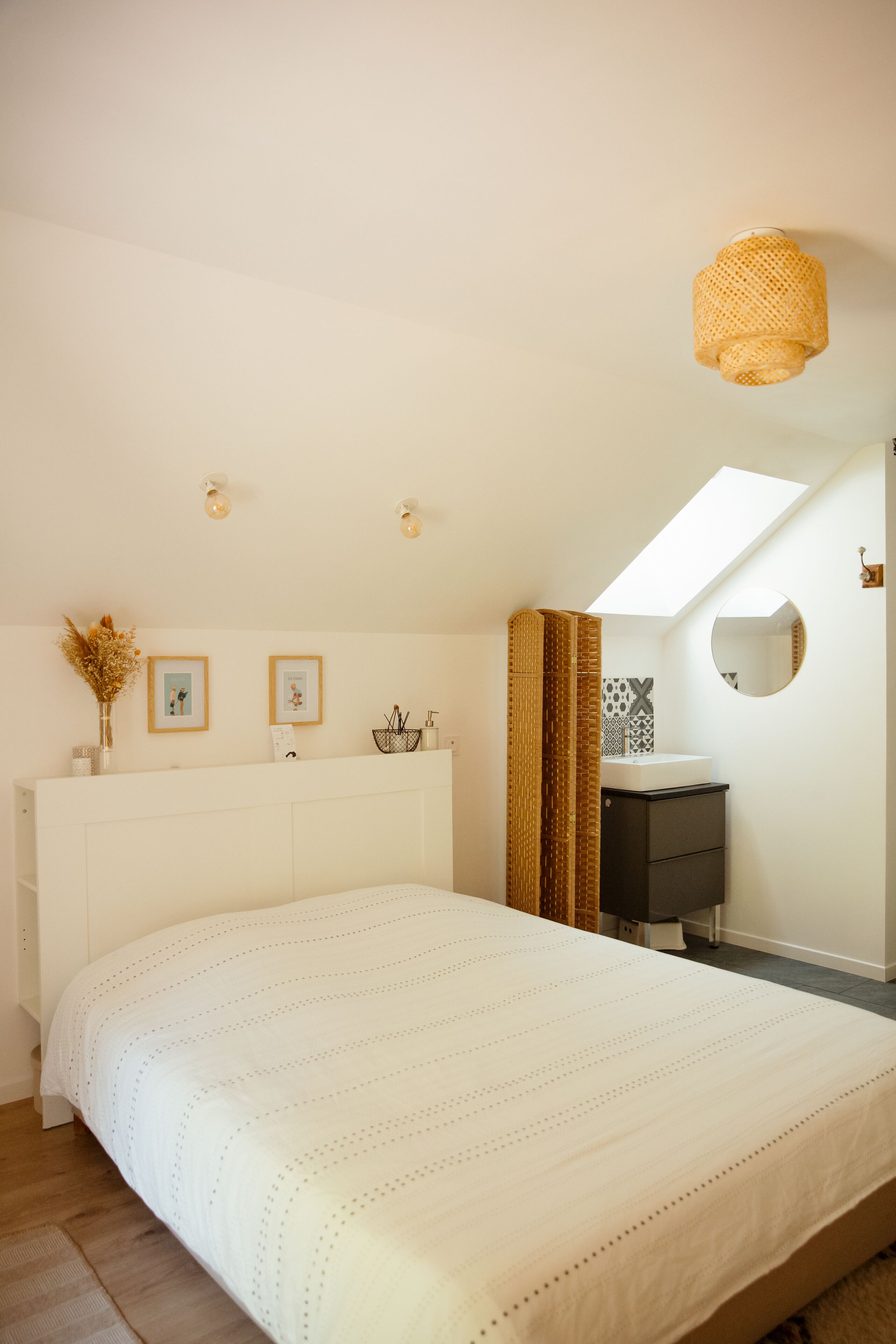 Een slaapkamer met een groot bed, dakraam, nachtkastje met spiegel en een kleine badkamer met wastafel en handdoekrek.