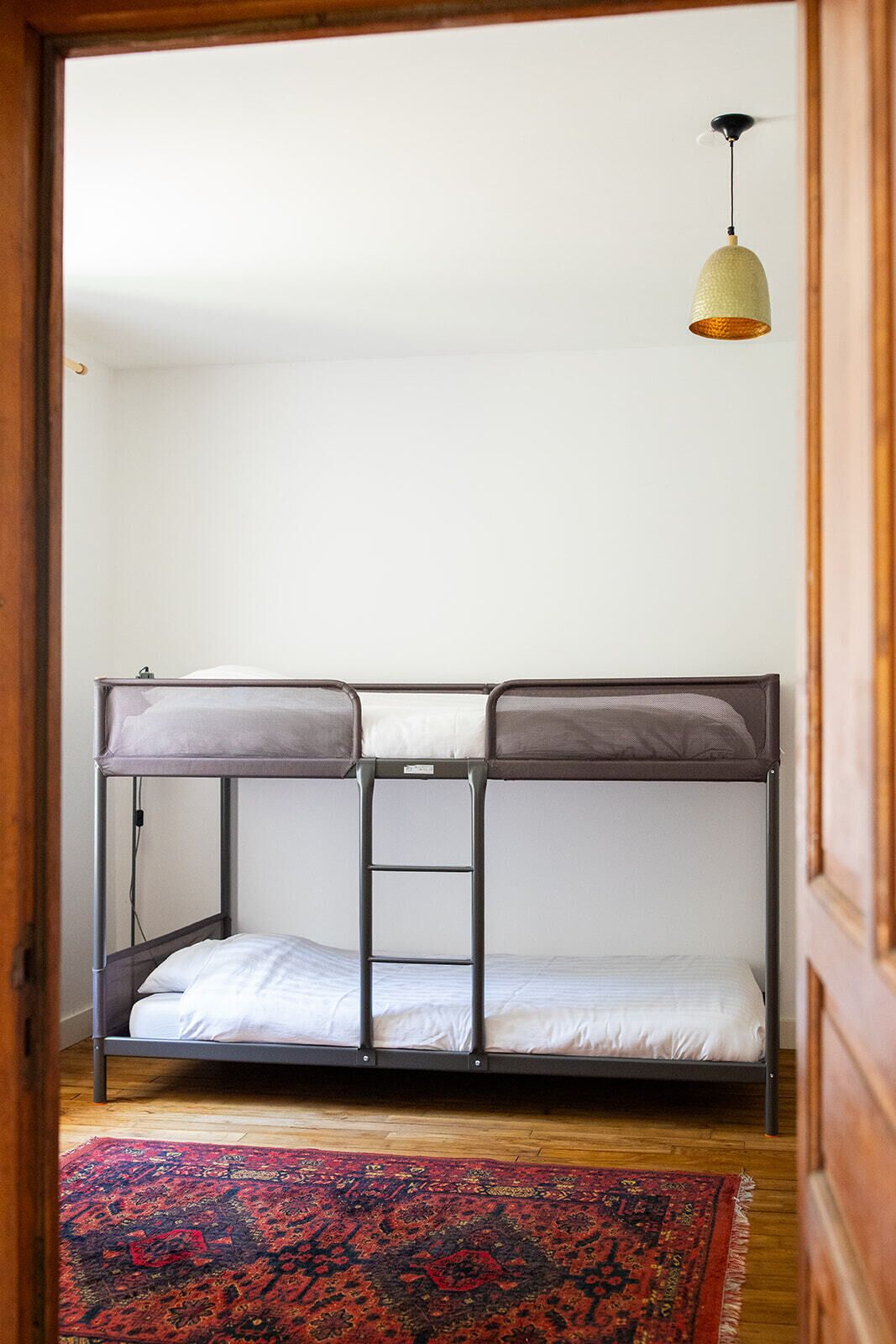 Een stapelbed met een ladder, een hanglamp en een getextureerd vloerkleed op een houten vloer.