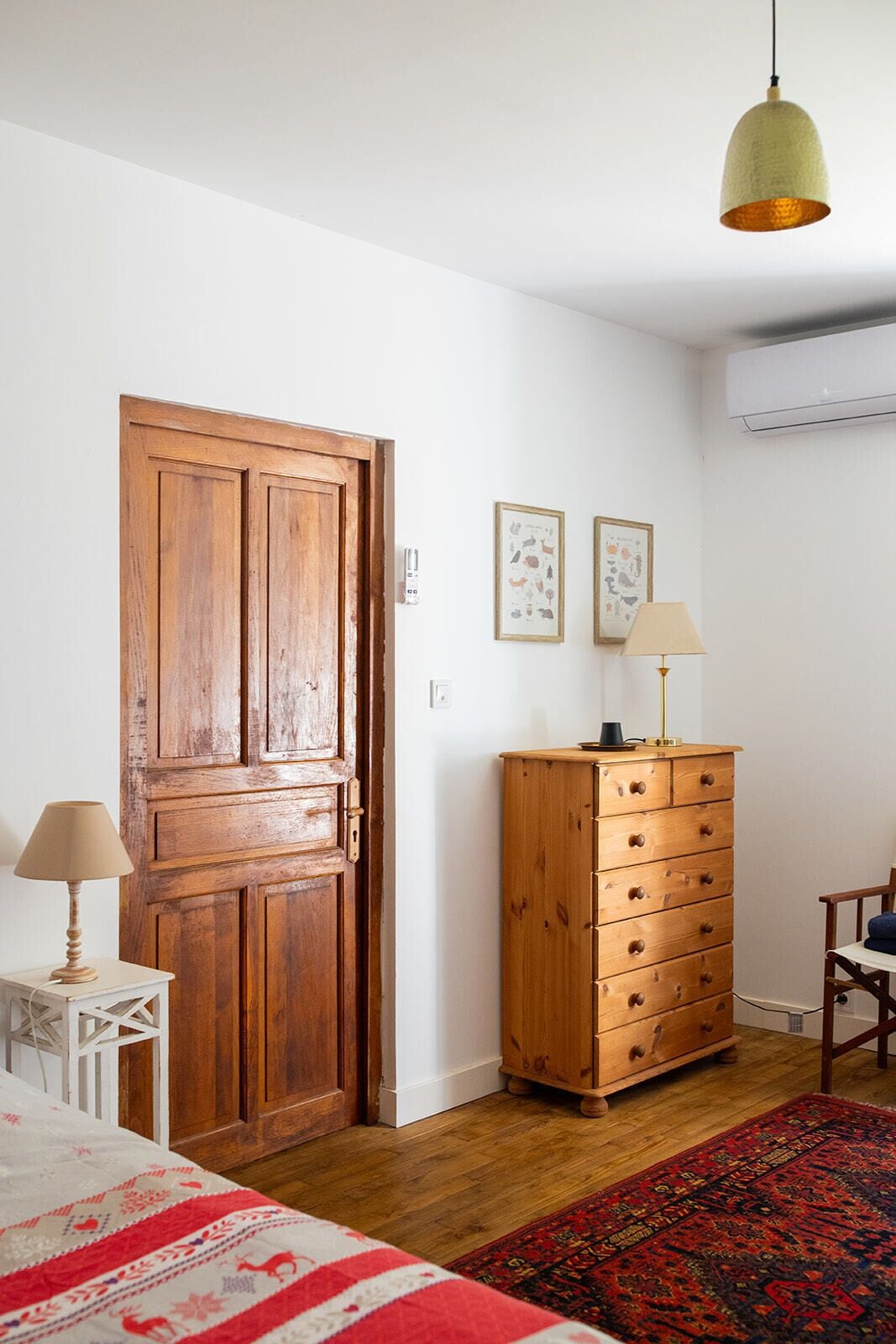 Una stanza con una porta in legno, un comò, una lampada, una sedia e un letto con copriletto a motivi rossi e bianchi.