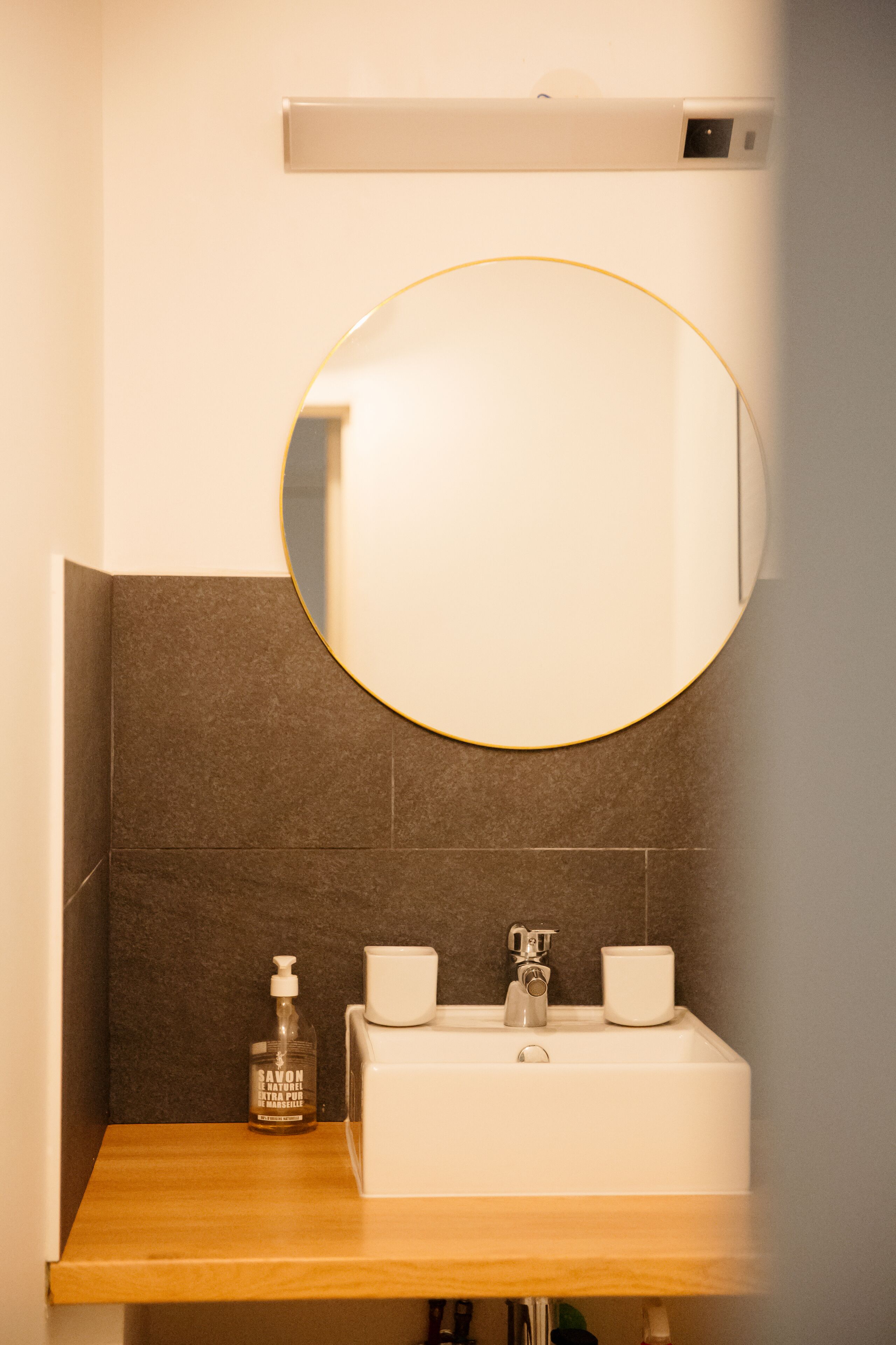A bathroom with a round mirror, a white sink, a soap dispenser, and a cup.