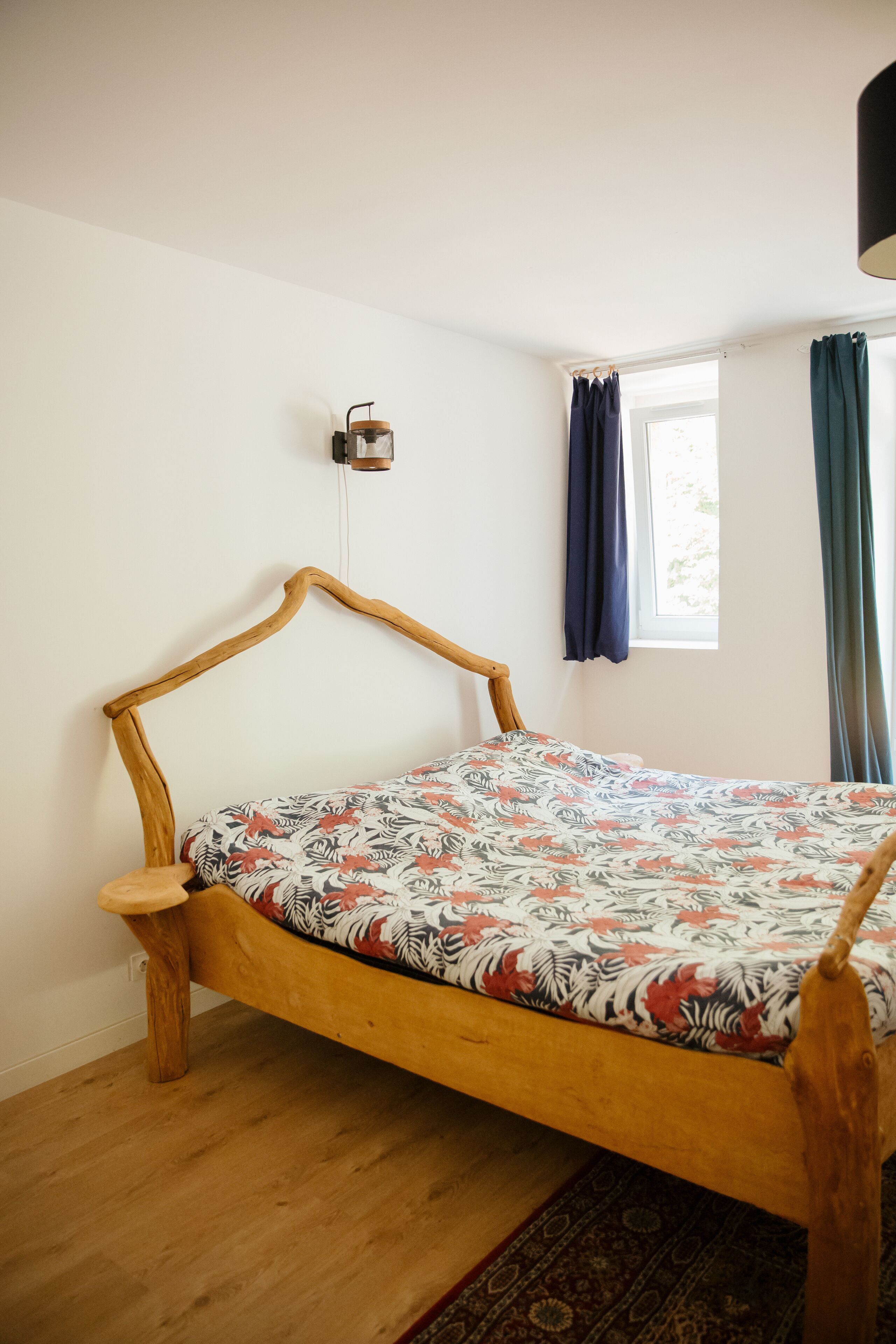 Un lit en bois recouvert d’une couvre-lit à motifs floraux, dans une pièce aux murs blancs et dotée d’une fenêtre avec des rideaux.