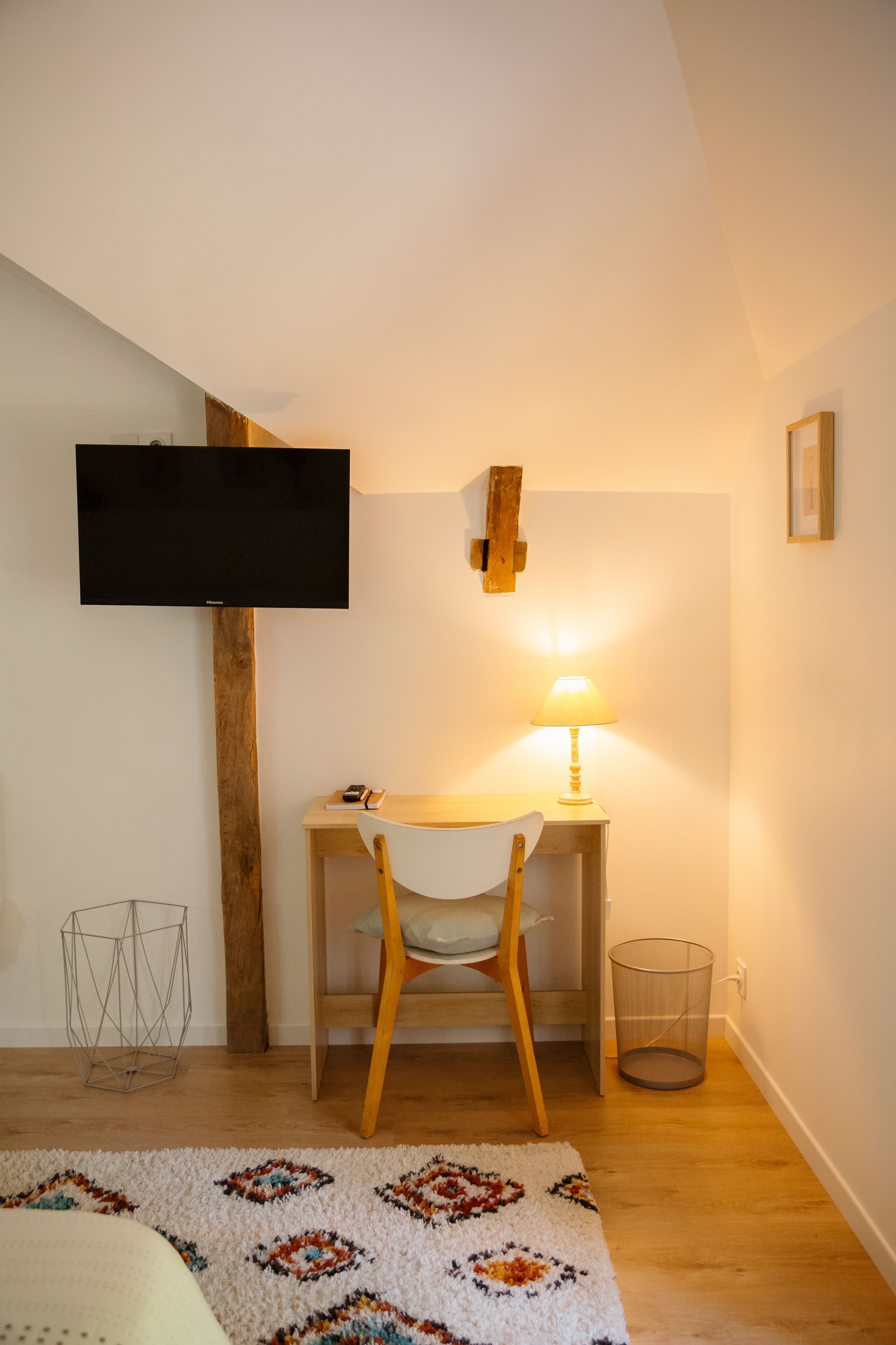 A bedroom with a bed, a desk with a chair, a TV mounted on the wall, a lamp, and a waste basket.