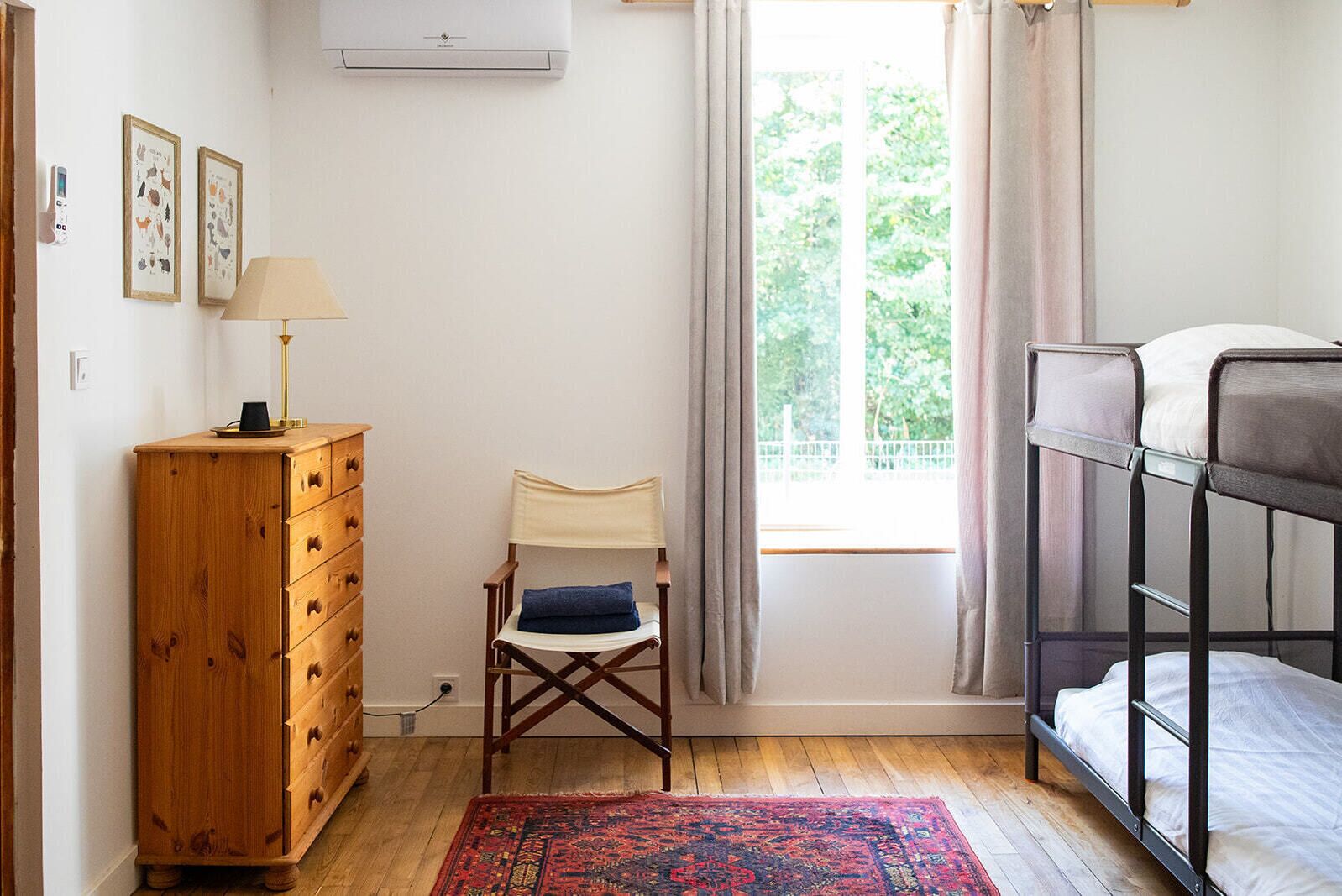 A room with a wooden dresser, a chair with a blue cushion, a bunk bed, a red patterned rug, and a window with curtains.