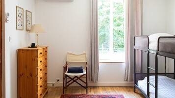 A room with a wooden dresser, a chair with a blue cushion, a bunk bed, a red patterned rug, and a window with curtains.