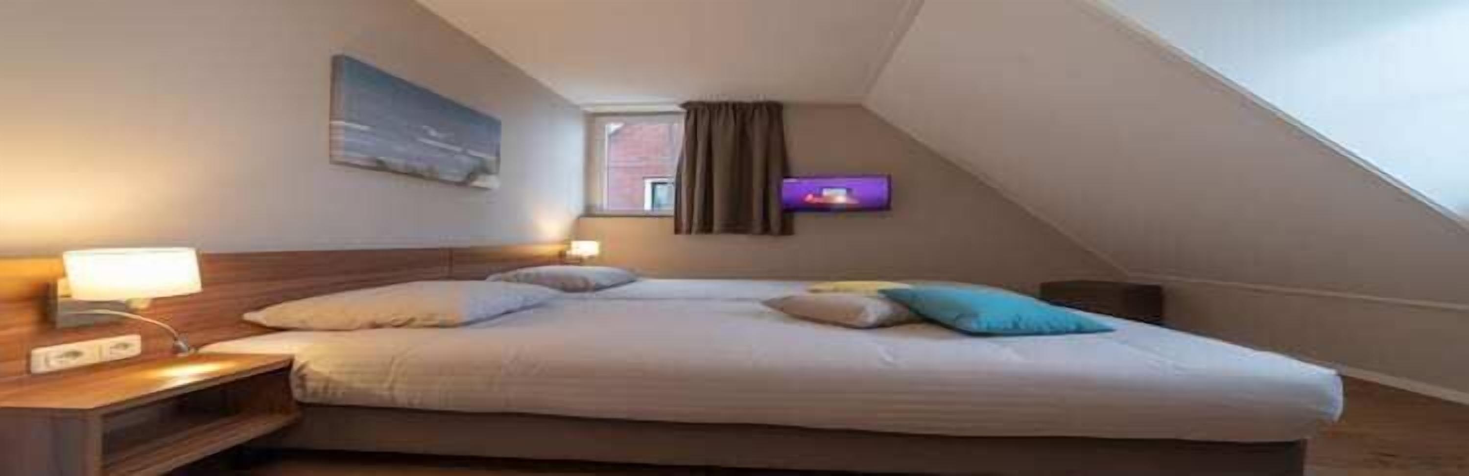 Ein Einzelbett in einem Zimmer mit schrägen Wänden, ein Nachttisch mit Lampe, ein Flachbildfernseher an der Wand und ein Gemälde über dem Bett.