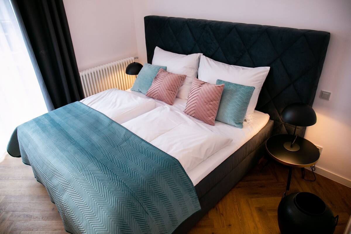 A bed with a dark headboard, multiple pillows, a teal blanket, a bedside table with a lamp, and a radiator.