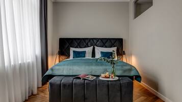 Un dormitorio moderno con cabecera capitonada, colcha color turquesa y un taburete oscuro con un libro y una vela.