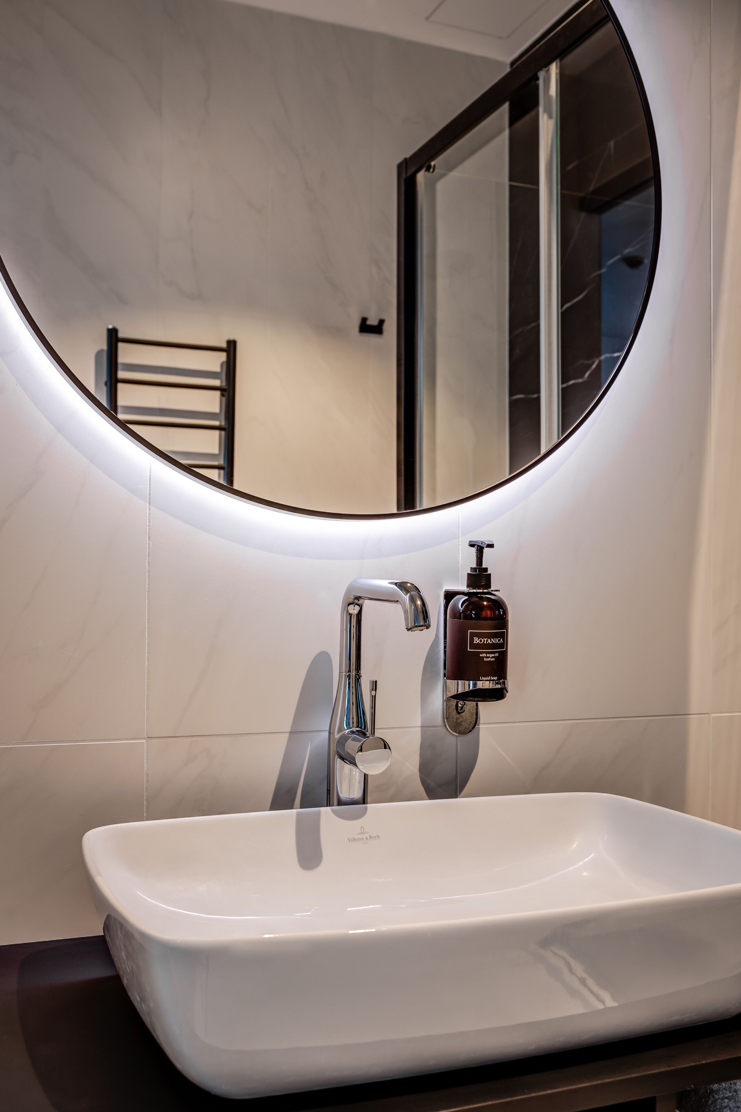 Une salle de bain avec un lavabo blanc, un miroir rond et un flacon de lotion.