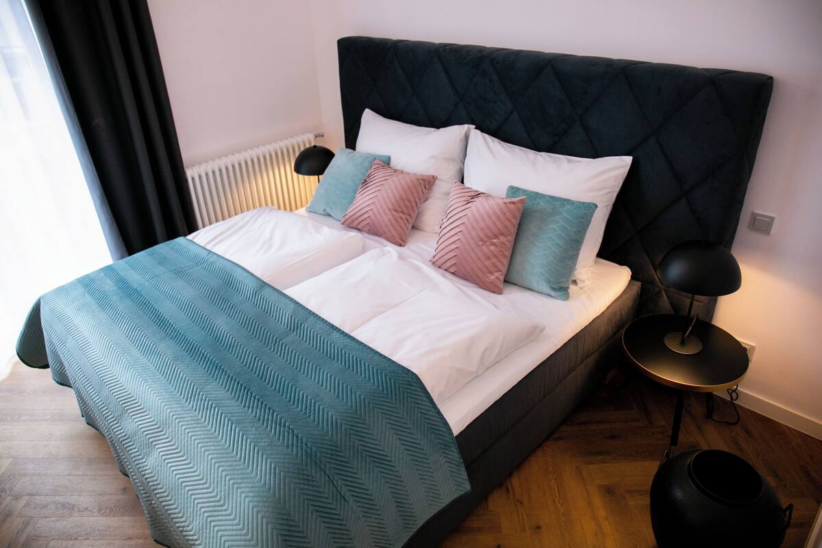 A neatly made bed with a quilted headboard, a bedside table with a lamp, and a radiator in the background.