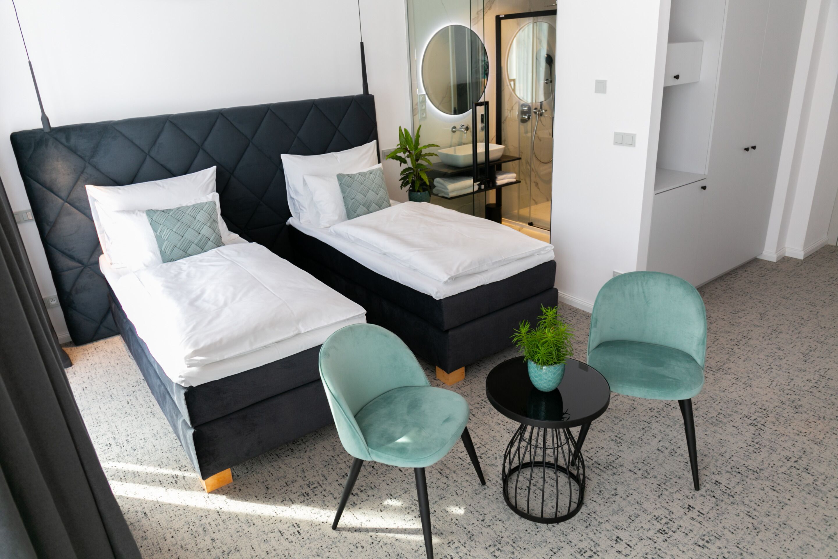 A modern hotel room with a black upholstered bed, two matching pillows, a small round table with a potted plant, and two teal chairs.
