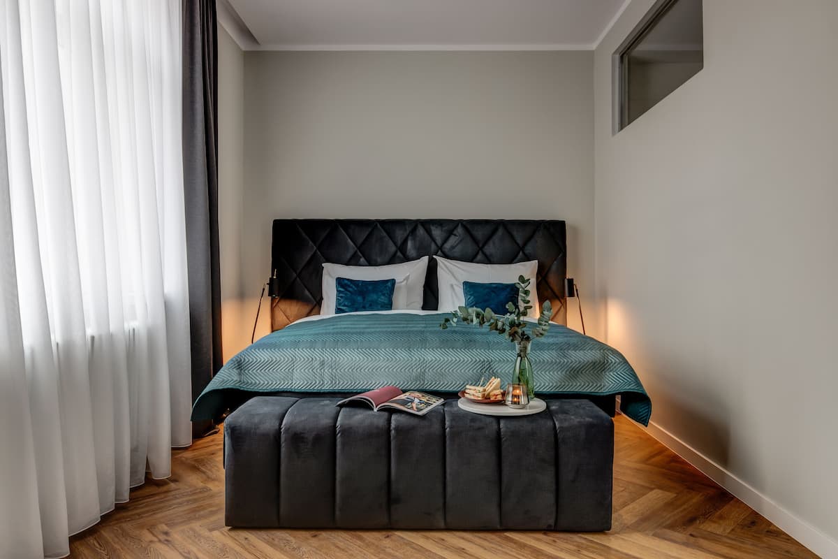 A bedroom with a bed, a bedside table, a window with curtains, and a wooden floor.
