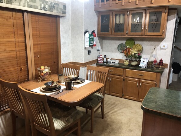 A dining area with a wooden table set for four, surrounded by wooden chairs. There are cabinets above the counter and a fire extinguisher mounted on the wall.