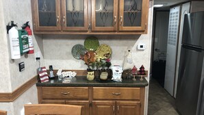 A kitchen with wooden cabinets, a stainless steel refrigerator, and a countertop with various items including a 'Home' sign, a vase with flowers, and a 'Faith Freedom' sign. - Halfway Tiny House - 3 BR Small Town Iowa Rural Setting! (Britt)
