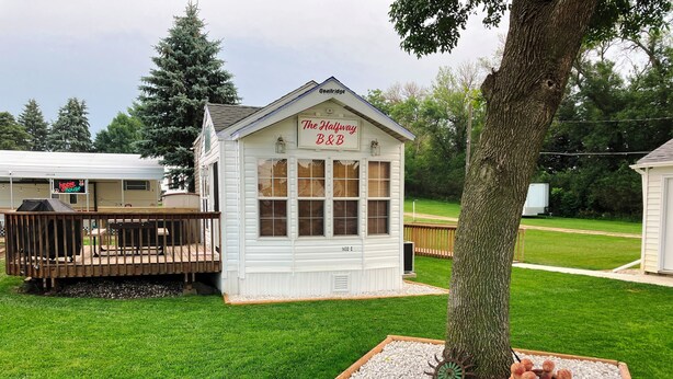 A small white building labeled "The Halfway B&B" with a deck and a tree in the foreground. - Halfway Tiny House - 3 BR Small Town Iowa Rural Setting! (Britt)