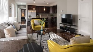 A modern living room with a sofa, yellow armchair, and a coffee table. There is a kitchen in the background with dark cabinets and a TV on a stand. - Stylish 2BR near it all (Cincinnati)