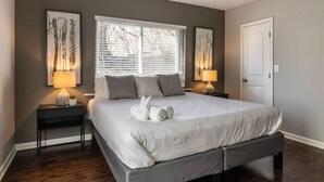 A bedroom with a large bed, two bedside tables with lamps, a window with blinds, and artwork on the walls. - Stylish 2BR near it all (Cincinnati)