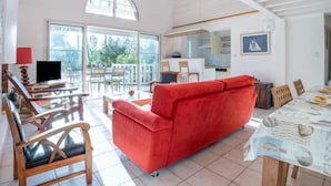 A spacious living area with a red sofa, wooden chairs, a dining table with a beach-themed tablecloth, and a kitchen in the background.