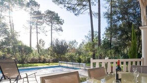 A poolside area with a glass table and chairs, a bottle of wine, and a view of the pool and surrounding trees.
