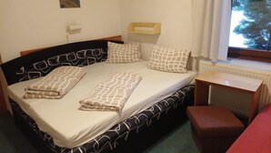 Ein Einzelbett mit gemusterten Betttextilien, ein Nachttisch, eine kleine Holzbank, eine Heizung und ein Fenster mit Blick auf Bäume.