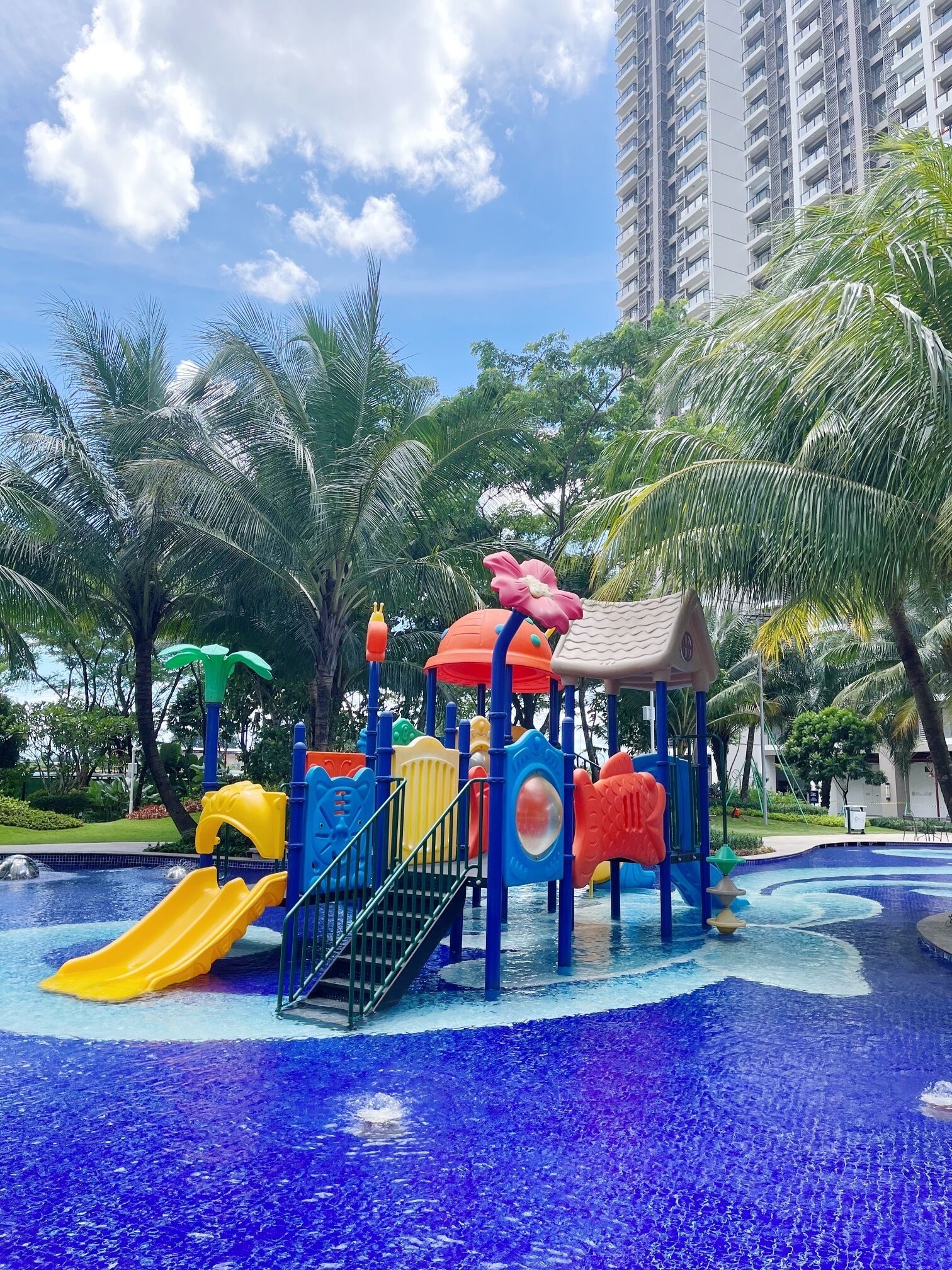 A children's play area with slides and climbing structures, surrounded by palm trees and a pool.