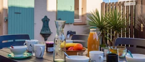 A wooden table set with white cups, saucers, a glass pitcher, a bottle of orange juice, and a bowl of fruit.