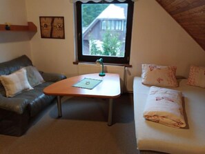 A cozy room with a brown leather sofa, a small wooden table with a lamp, a white sofa with floral cushions, and a window with a view of greenery outside.