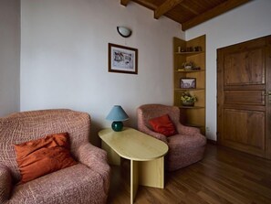 Ein Zimmer mit zwei gemusterten Sesseln, einem kleinen Holztisch, einer Lampe, einem gerahmten Bild an der Wand und einer Holztür.