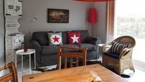 Een woonkamer met een grijze bank, twee rode kussens met witte sterren, een houten eettafel, een rieten stoel, een rode vloerlamp en een aan de muur hangend schilderij.