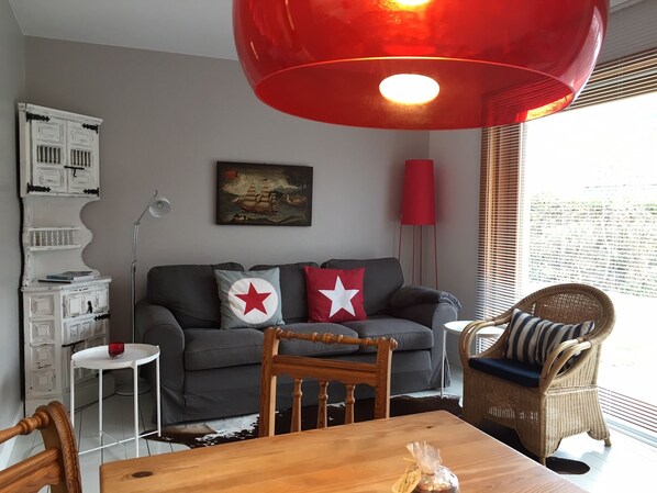 A living room with a grey sofa, two red pillows with white stars, a wooden dining table, a wicker chair, a red floor lamp, and a wall-mounted painting.