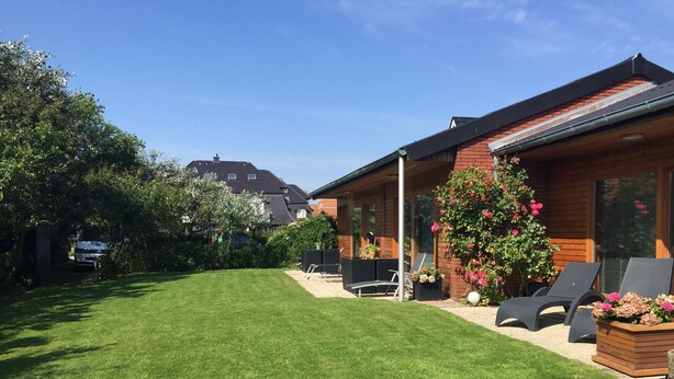 Een verzorgd gazon, een houten huis met terras, en een tuin vol bloemen.