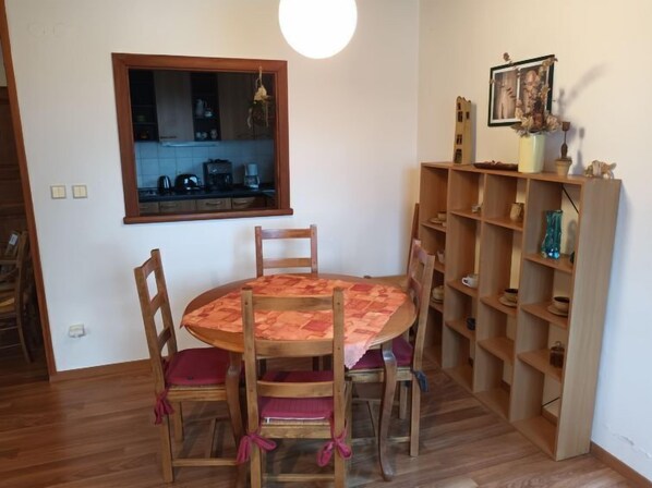 A dining area with a round table, four wooden chairs, a wooden bookshelf, and a kitchen visible through a window.