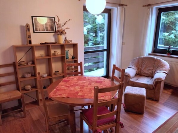 A cozy room with a wooden dining table, chairs, a bookshelf, and a window with a view of greenery.