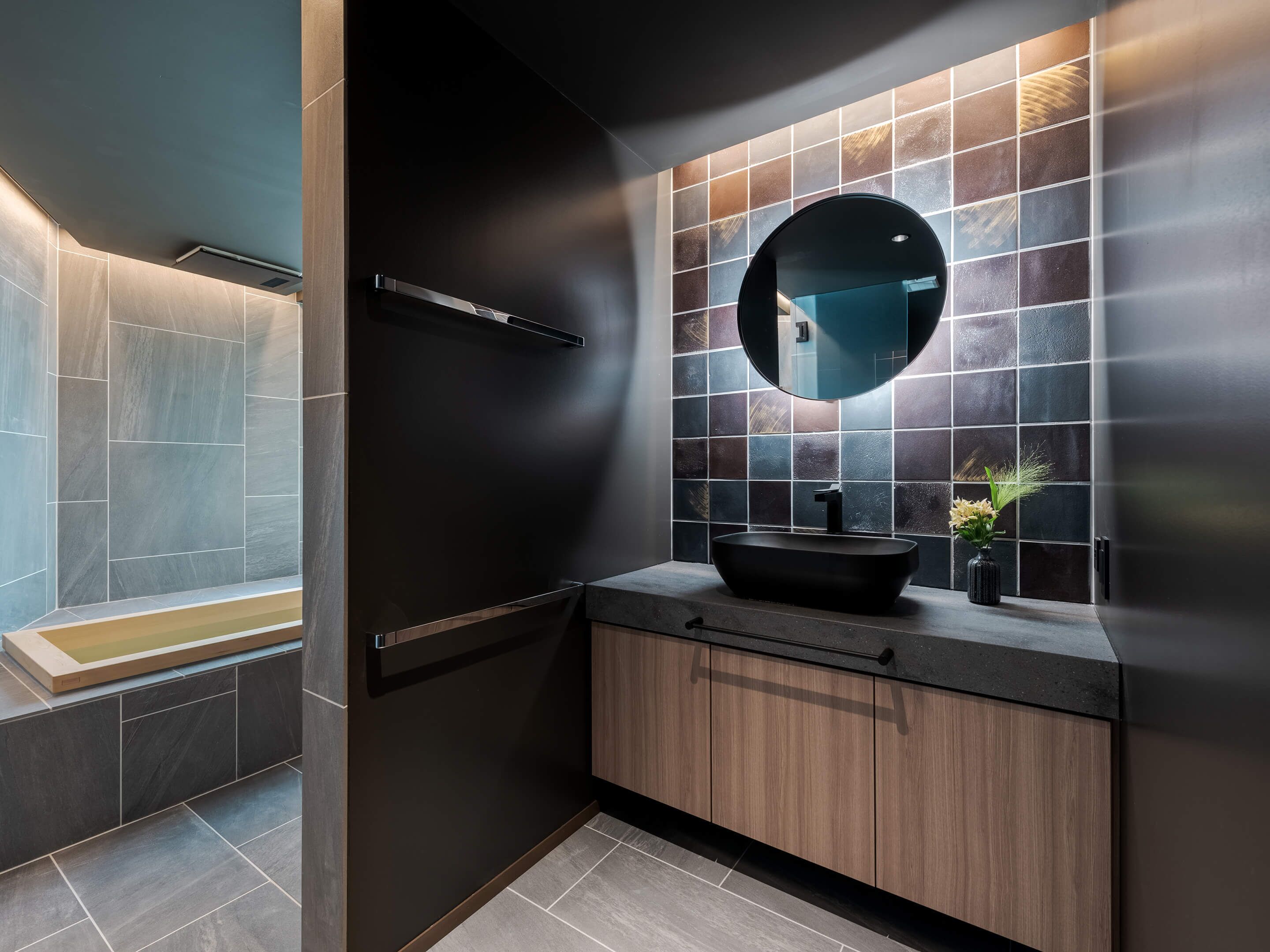 Een moderne badkamer met een zwart wastafelmeubel, een ronde spiegel en een betegelde douchehoek.