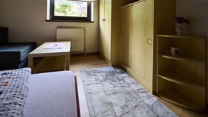 Una habitación pequeña y luminosa con una cama, una mesa, un radiador, un ventanal con persianas y un armario de madera.