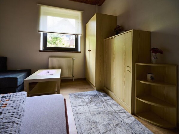 Une petite pièce bien éclairée, avec un lit, une table, un radiateur, une fenêtre avec des stores et une armoire en bois.