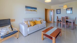 A living room with a sofa, a wooden coffee table, a dining area with a table and chairs, and a door leading to another room. - Vista Mar by Interhome (Albufeira)
