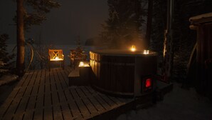 A wooden deck with a hot tub and seating area, surrounded by snow-covered trees.