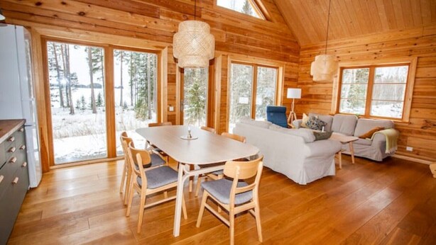 A log cabin interior with a dining table, chairs, a sofa, and a large window overlooking a snowy landscape.