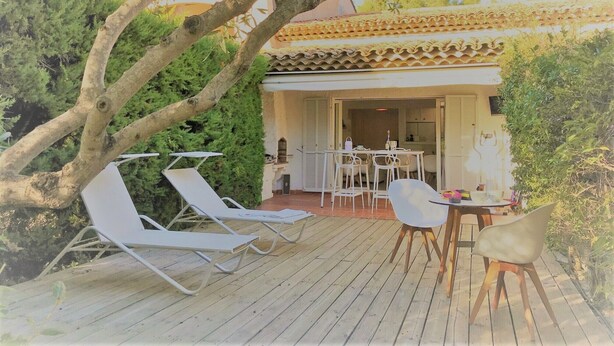 Eine Holzterrasse mit weißen Liegestühlen und einem Tisch, umgeben von viel Grün und einem Gebäude mit Ziegeldach.