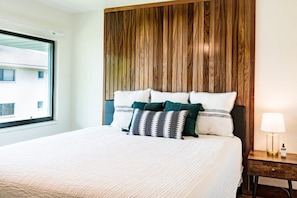 Un lit bien fait, avec une couette blanche et plusieurs oreillers, une tête de lit en bois, une table de chevet avec une lampe et une fenêtre donnant sur des immeubles.