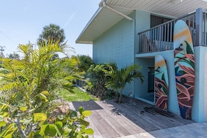 Une maison à deux étages dotée d’une terrasse en bois, entourée de plantes tropicales et ornée d’une fresque colorée représentant une planche de surf.